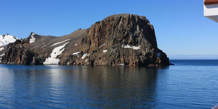Blick von einem Schiff auf einen schroffen Berg, der aus der arktischen See ragt. 
