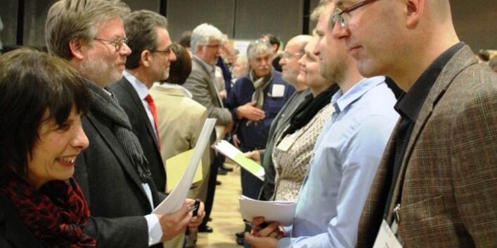Zahlreiche Personen stehen sich in zwei Schlangen gegenüber. Immer zwei Personen schauen sich an und reden miteinander. Sie grinsen, wirken aufgeregt und sind schick gekleidet.
