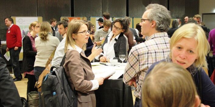 Die Teilnehmenden stehen verteilt um Stehtische und tauschen sich angeregt aus. Im Vordergrund steht eine blonde Frau und in der Mitte eine Frau mit einem schwarzen Rucksack, die mit einem Herren im karierten Hemd spricht. Hinter ihnen sind noch sehr viele weitere Personen zu sehen.