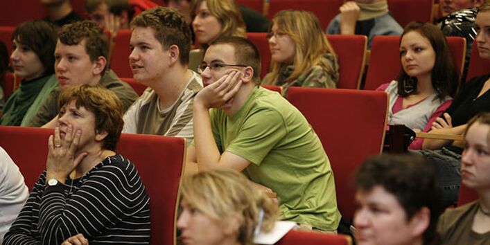 Blick auf das Publikum. Ein Schüler und eine Lehrerin hören interessiert zu, das Gesicht in die Hände gestützt.