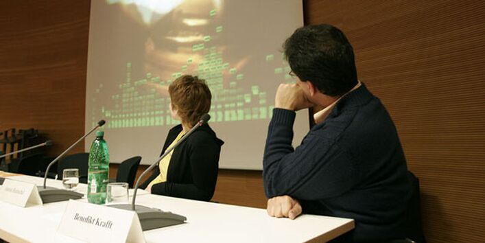 Zwei Podiumsdiskutanten blicken auf eine Leinwand hinter sich, auf der ein Video gezeigt wird.