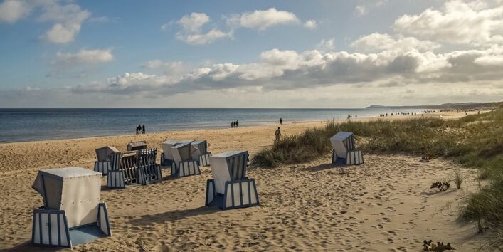 The picture shows a sandy beach on the Baltic Sea coast with dunes and hooded beach chairs (‘strandkorbs’). The strandkorbs facing the viewer have been locked up for the winter. The sea is visible on the horizon. A few individuals and small groups of people are visible at the waterline; apart from them, the beach is deserted. The sky is partly cloud-covered. 