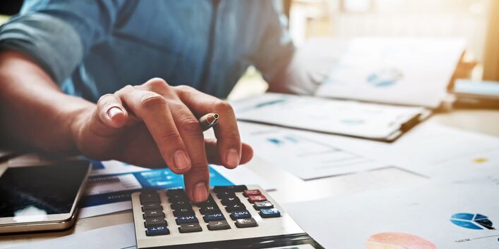 The picture shows a man sitting at a desk holding a ballpoint pen between his fingers while operating a pocket calculator. There are several documents on the desk; it is just possible to make out the fuzzy image of some charts. 