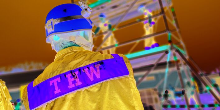 Das Bild zeigt einen Mitarbeiter des Technischen Hilfswerks in Uniform und mit Helm von hinten. Im Hintergrund ist ein Gerüst zu erkennen, auf dem weitere Mitarbeitende des THW stehen.