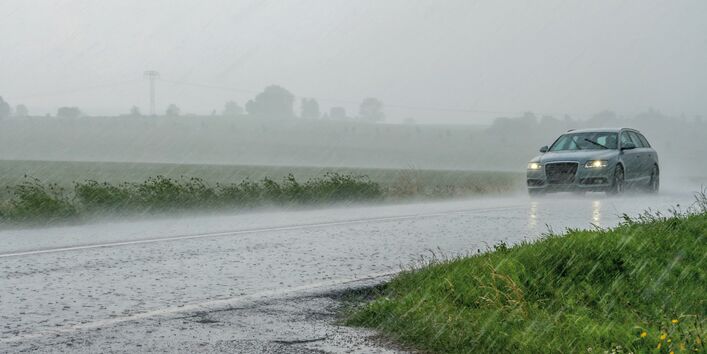 The picture shows a car driving on a road covered in water. It is clear to see that the headlights and windscreen wipers are switched on. Heavy rain is falling.