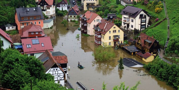 The picture shows a village – situated on the slope of a hill – inundated by floodwater. Some of the lower buildings and some lower floors of houses have been flooded.