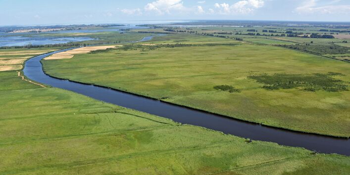 The picture shows a river running through a flat landscape consisting of meadows and agricultural areas. The sea is visible in the background.
