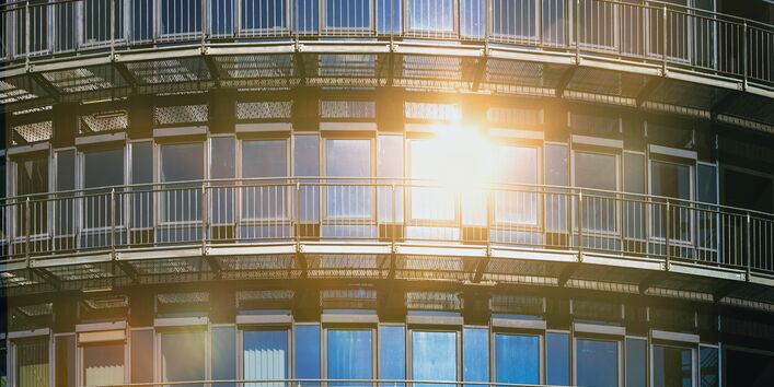 Das Bild zeigt die Glasfront eines Hauses, in der sich die Sonne spiegelt. Einige Fenster sind mit Außenrollos teil-verschattet.