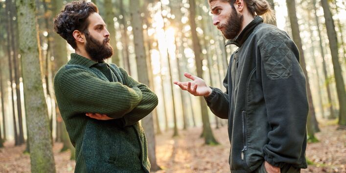 The picture shows two young foresters having a discussion in a sparse forest. The man on the left-hand side of the picture stands with his arms crossed while the other man is gesticulating with his hands. In the background, beams from a low sun are penetrating the forest.