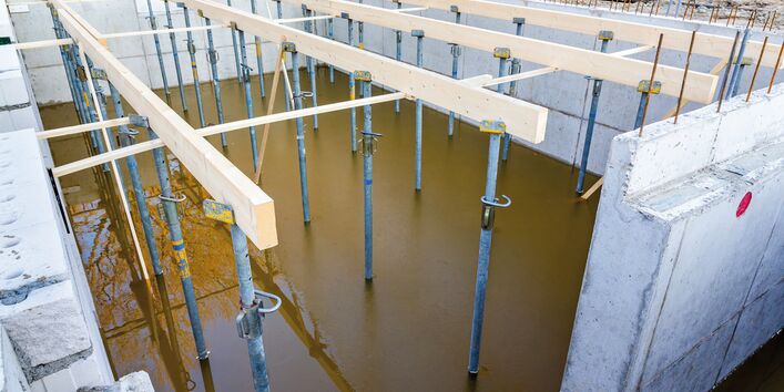 The picture shows a building pit where the walls of a cellar have already been built. Steel girders are seen supporting wooden beams which extend across the ceiling of the cellar. The bottom of the building pit is filled with brown muddy water reflecting a tree.