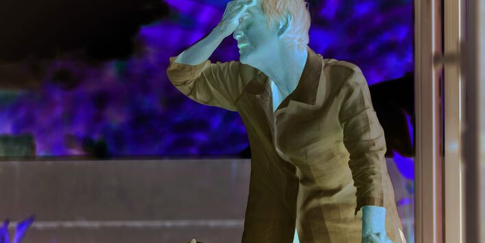 The picture shows an elderly woman wearing a white blouse. The lady is holding on to a rollator with one hand, while she is holding the back of her left hand to her forehead. In the background, a few fuzzy treetops or bushes are visible in bright light.