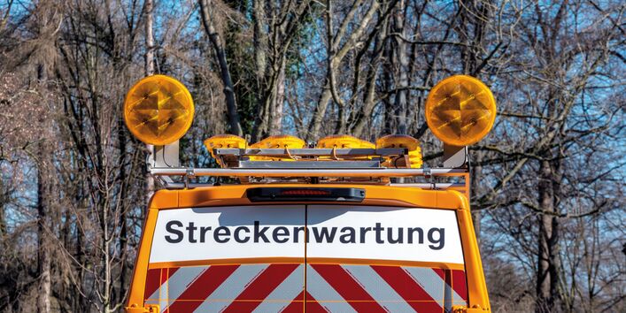 The picture shows the upper part of the tail end of a vehicle belonging to the highway maintenance service. The vehicle is orange and its rear doors are marked with the word ’Streckenwartung’ (track maintenance) and reflective red-and-white striped stickers. There are warning lights fitted to the vehicle.