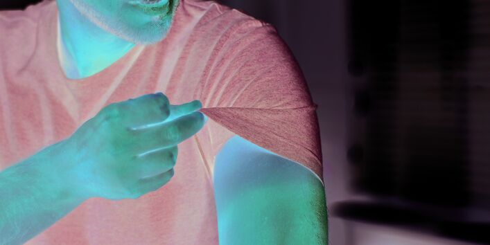 The picture shows a man’s upper arm in focus. The man is lifting the sleeve of his T-shirt with two fingers. In contrast with the skin previously covered by the T-shirt, the part of the skin that has been exposed to sunlight looks very red.