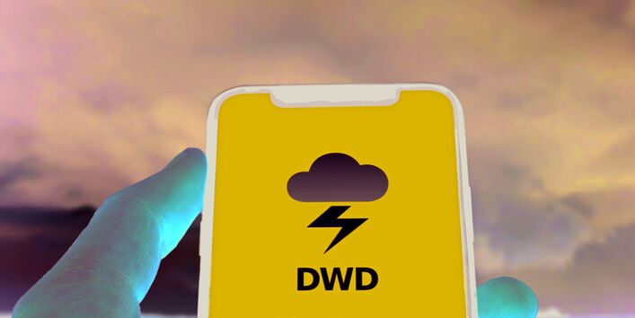 Das Bild zeigt eine Hand, die ein Smartphone hält. Der Bildschirm des Smartphones ist blau und zeigt das Logo der WarnWetter-App des DWD. Im Hintergrund des BIldes sind die schwarzen Wolken eines Gewitterhimmels zu erkennen.