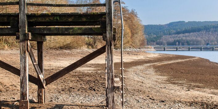 Das Bild zeigt einen Steg, der im ausgetrockneten sandig-steinigen Uferbereich eines Sees steht. Im Hintergrund ist eine Brücke und Restwasser zu sehen.