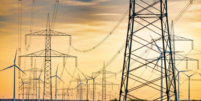 The picture shows numerous high-voltage pylons and power lines against a cloudy, yellowish sky. A few modern wind turbines are visible in the background. 