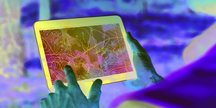 Das Bild zeigt Hände, die ein Tablet halten, auf dessen Bildschirm eine Karte mit Höhenlinien und weiteren Markierungen zu sehen ist. Der verschwommene Bildhintergrund zeigt, dass die Person, die das Tablet hält, in einem Wald steht.