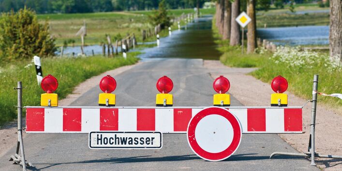 The picture shows a flooded dip in a road. There is a roadblock in the foreground indicating that the road is closed to traffic. A sign reading ‘Hochwasser’ (floodwater) is fixed to the roadblock. There are flooded meadows visible on both sides of the road.  