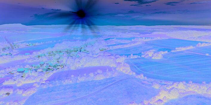 The picture shows a slightly hilly landscape characterised by agricultural activity. The centre of the picture shows a village embedded in the landscape. In an almost cloudless sky which is already tinged with orange, you can see a low sun wrapping the landscape in a warm light.