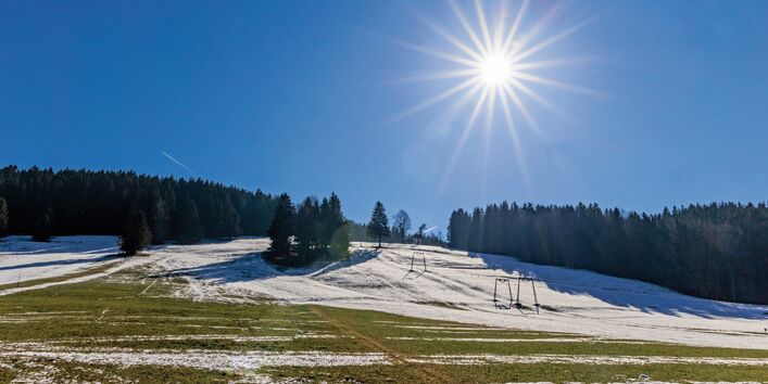 The picture shows a ski lift heading for a slope wooded predominantly with conifer trees. The slope is covered with a thin layer of snow. However, in the foreground especially, there are large areas of ground without any snow cover thus revealing grass and bare earth. A radiant sun is shining brightly from a cloudless sky.