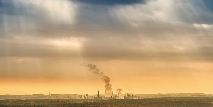 Das Bild zeigt einen bewaldeten Landschaftsausschnitt. In der Ferne ist ein aus mehreren Gebäuden und Anlagen bestehendes Industriegebiet zu sehen. Aus einigen Schloten steigt Rauch empor. Der Himmel ist stark bewölkt. Am Horizont ist er gelblich gefärbt, am oberen Rand des Bildes von dunklen, grau-blauen Wolken verhangen. An wenigen lichteren Stellen dringen Sonnenstrahlen durch das Wolkenband.