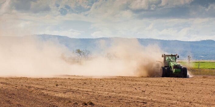 The picture shows a dry-looking arable field with a tractor being driven across it. Owing to drought, the vehicle is stirring up clouds of dust. In the background, it is possible to make out a ridge of hills. 