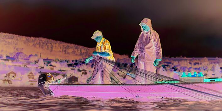Das Bild zeigt zwei Männer in wasserfester Arbeitskleidung, die auf dem Bodensee in einem kleinen Boot stehen. Einer von ihnen hält ein Fischernetz in den Händen. Er scheint es aus dem Wasser zu ziehen. Im Hintergrund ist das Ufer mit einer Siedlung zu erkennen.