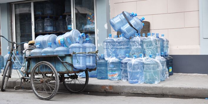 Altes Lastendreirad, welches auf der Straße abgestellt wurde und mit leeren Wasserkanistern vollbepackt ist; auf dem Fußgängerweg lagern weitere leere Plastikkanister; einige sind in Plastiktüten eingepackt, andere nicht; die Kanister kommen aus einem Lager, welches offen ist