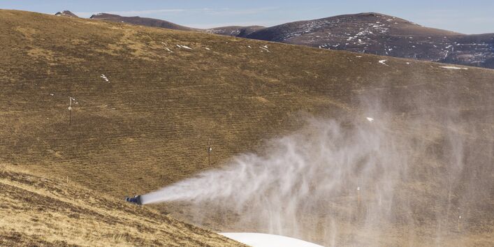 The picture shows an operating snow cannon in a mountainous slope area where there is no snow.