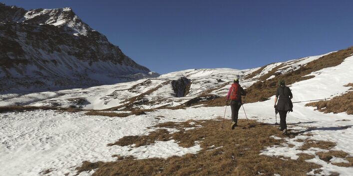 Das Bild zeigt zwei Bergsteigerinnen von hinten, die mit Stöcken über eine teilweise schneebedeckte Bergwiese wandern.  