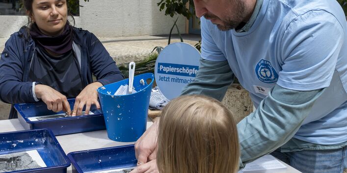 Gäste und Standbetreuer mit Altpapier, Wasser und Formen mit dem Umweltzeichen Blauer Engel, um daraus einen Engel zu schöpfen. 