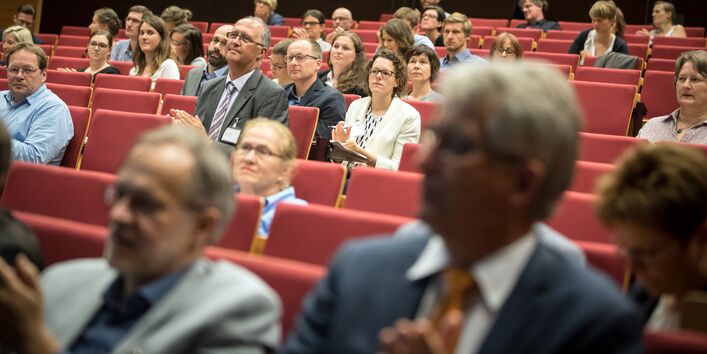 Blick von vorne in die Sitzreihen des Hörsaales. Veranstaltungsteilnehmerinnen und -nehmer sitzen zuhörend auf roten Reihenstühlen.
