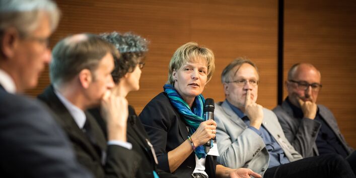 Personen sitzen zur Interviewrunde. Eine Teilnehmerin hält das Mikrofon in der Hand und spricht hinein.