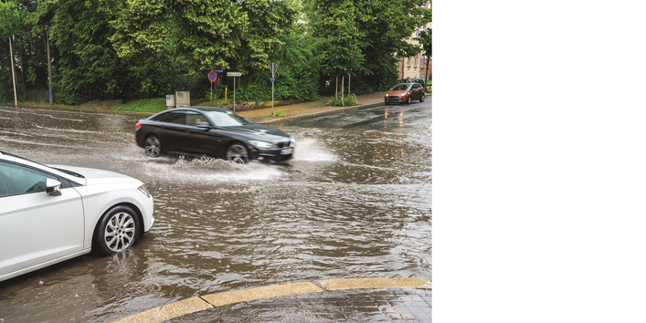 Das Bild zeigt eine völlig überflutete Straßenkreuzung in einer Stadt, in die gerade zwei Autos hineinfahren.