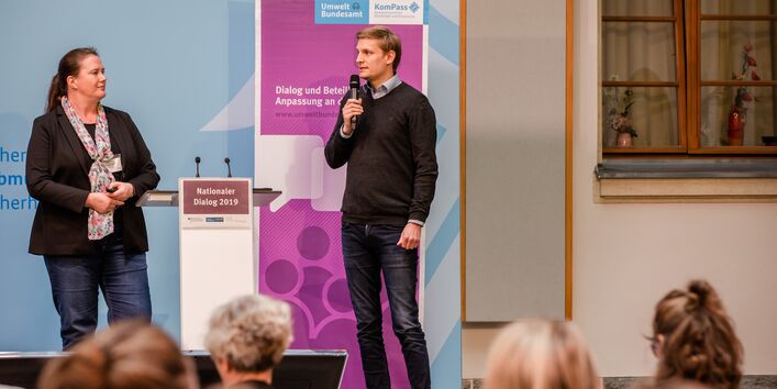 Zwei Teilnehmende stehen am Stehpult vor dem blauen Hintergrund, auf dem der Veranstaltungsort geschrieben steht sowie das lilafarbene Veranstaltungs-Rollup steht.