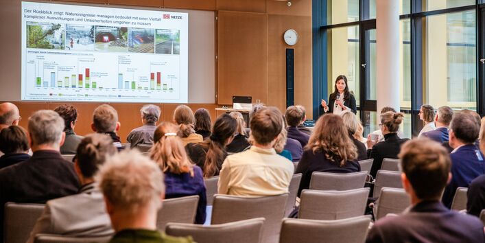 Teilnehmende folgen auf beigen Stühlen in Reihen sitzend einer Präsentation. Die Vortragende steht mit Handmikrofon nahe Stehpult vor einer Fensterfront.