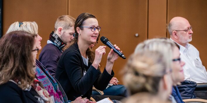Eine Teilnehmerin spricht angeregt ins Handmikrofon. Alle Teilnehmende sitzen in Reihen im Veranstaltungsraum. Die Wände sind holzvertäfelt.