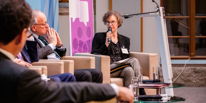 Podiumsdiskussion aus einer anderen Perspektive. Im Hintergrund steht ein Rollup in lila Farbtönen mit Veranstaltungstitel. Die Moderatorin spricht in das Mikrofon.