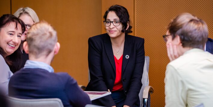 Teilnehmende sitzen in aufgelockerter Stimmung beieinander im holzfarben vertäfelten Veranstaltungsraum. Die Teilnehmerin im Mittelpunkt trägt ein dunkelblaues Sacko mit rotes Oberteil.