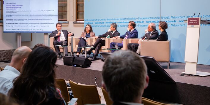 Podiumsdiskussion aus einer anderen Perspektive. Im Hintergrund steht eine Tafel, auf der die Podiumsbeteiligten verzeichnet sind.