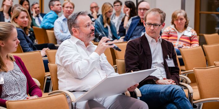 Teilnehmende sitzen im einem Veranstaltungsraum auf orangenen Stühlen in Reihen und blicken nachdenklich. Ein Teilnehmer spricht in ein Handmikro und hält ein Tableau in der Hand.