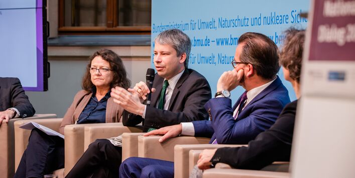 Podiumsdiskussion vor hellblauem Hintergrund, auf dem der Veranstaltungsort geschrieben steht.