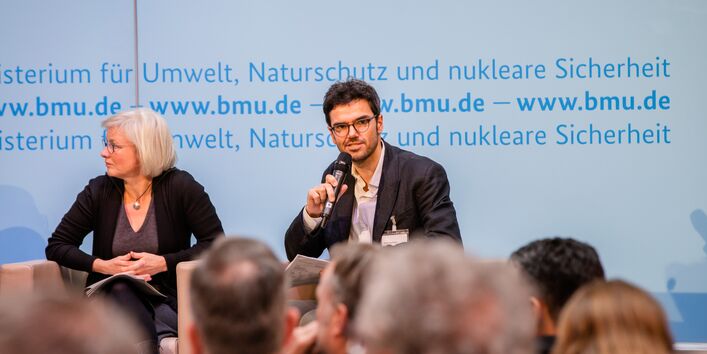 Podiumsdiskussion vor dem hellblauem Hintergrund, auf dem der Veranstaltungsort geschrieben steht. Ein Teilnehmender hält ein Handmikrofon.