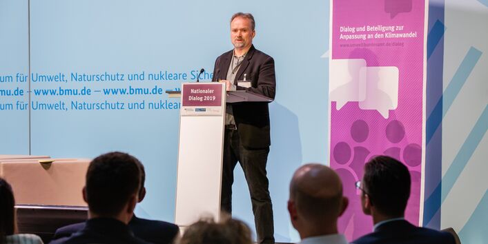 Vortragender steht am Stehpult vor blauem Hintergrund, auf dem der Veranstaltungsort geschrieben steht. Außerdem steht daneben das in lila Farbtönen gehaltene Veranstaltungs-Rollup.