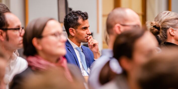 Der Blick geht durch die Teilnehmenden auf einen Teilnehmer, der sehr nachdenklich schaut. Er trägt einen blauen Anzug mit hellblauem Schlips und weisses Hemd. Der Raum ist hell und holzfarben abgesetzt.