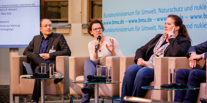 Podiumsdiskussion vor dem hellblauem Hintergrund, auf dem der Veranstaltungsort geschrieben steht. Eine Teilnehmende hält ein Handmikrofon und spricht angeregt zu den anderen Podiumstteilnehmenden.