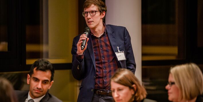 Teilnehmender steht im Veranstaltungsraum und spricht in ein Mikrofon, welches er in seiner rechten Hand hält.