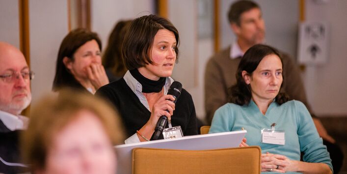 Teilnehmende sitzen im einem Veranstaltungsraum auf orangenen Stühlen in Reihen und blicken nachdenklich. Eine Teilnehmerin spricht in ein Handmikro.
