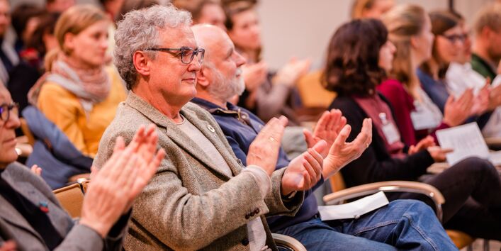 Teilnehmende sitzen auf orangenen Stühlen in Reihen im Veranstaltungsraum und applaudieren.