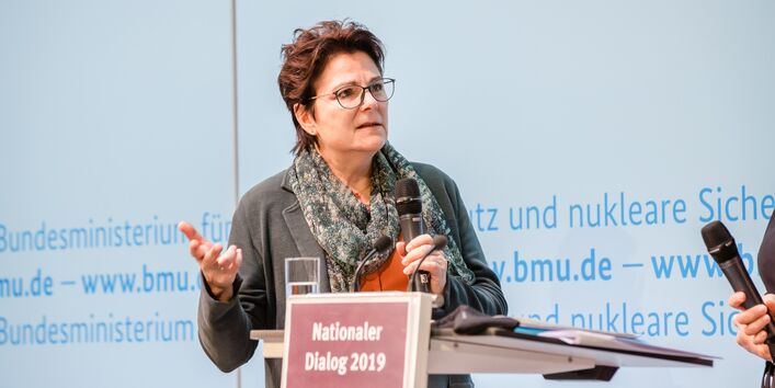 Zwei Vortragende stehen am Stehpult vor blauem Hintergrund, auf dem der Veranstaltungsort geschrieben steht. Beide halten Handmikrofone in den Händen und sprechen gestikulierend  miteinander.